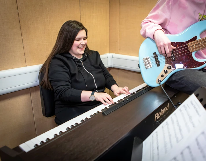 A smiling student in a black hoodie plays a Roland keyboard while another plays a blue bass guitar beside them. A smiling student in a black hoodie plays a Roland keyboard while another plays a blue bass guitar beside them.